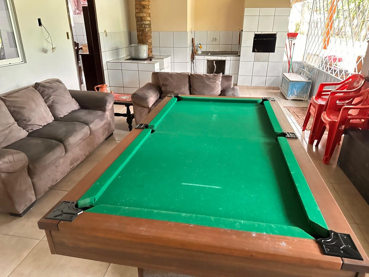 An area featuring a green felt pool table as the focal point, surrounded by a comfortable sofa set and red plastic chairs. Light neutral walls complement the layout, while a fireplace is visible in the background, enhancing the communal feel of the space.