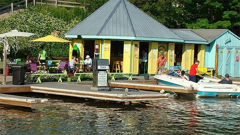 A colorful, waterfront building with a yellow and blue facade is visible. Various outdoor seating areas are arranged under umbrellas, with patrons enjoying their time. Two small boats are docked at the wooden slip, while gentle ripples in the water reflect the surrounding greenery.