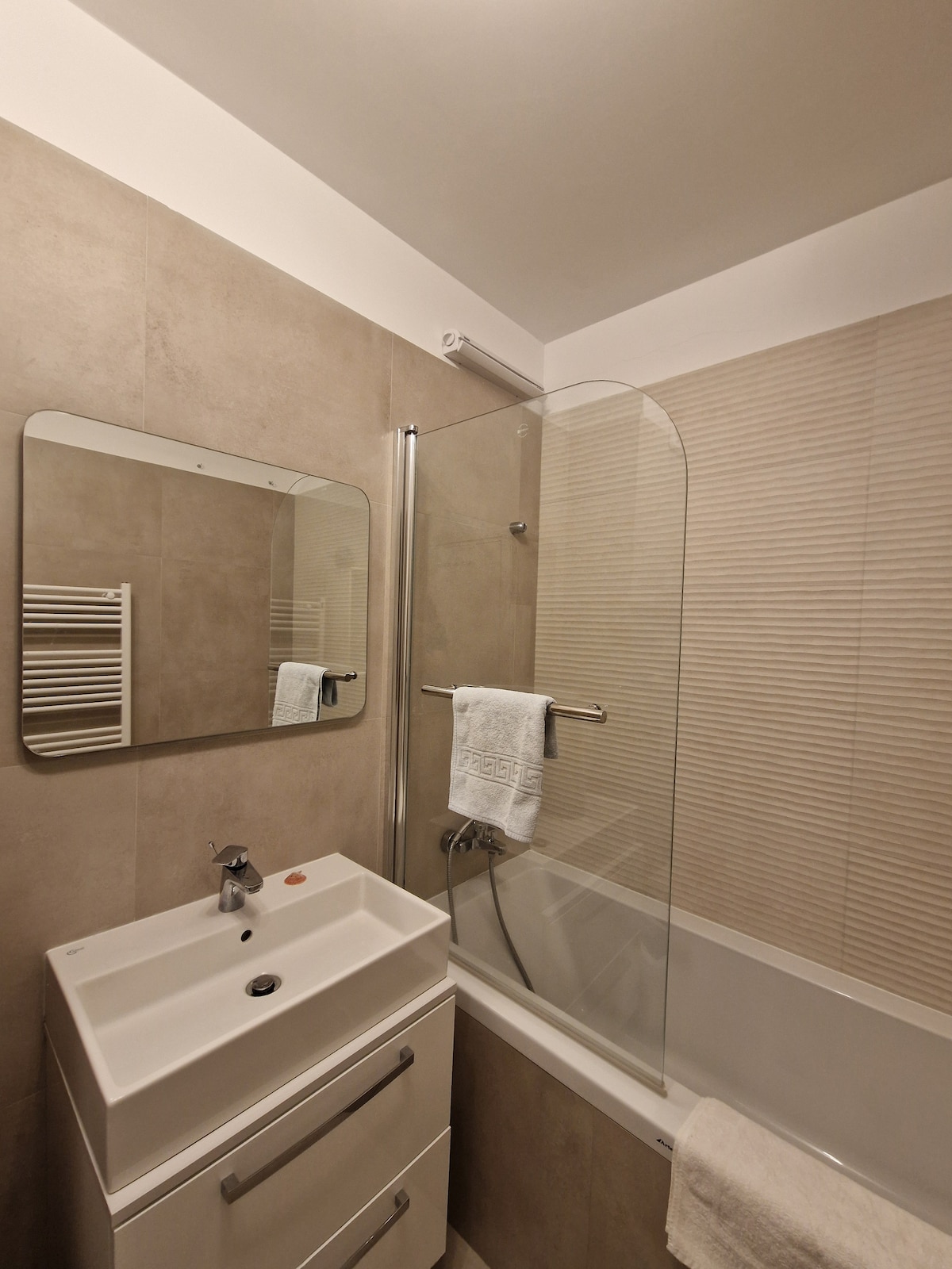 A modern bathroom features clean lines and neutral tones. A glass shower screen separates the shower area from a bathtub. A compact vanity with a sleek sink is complemented by a large mirror, while towels hang neatly on a heated towel rail.