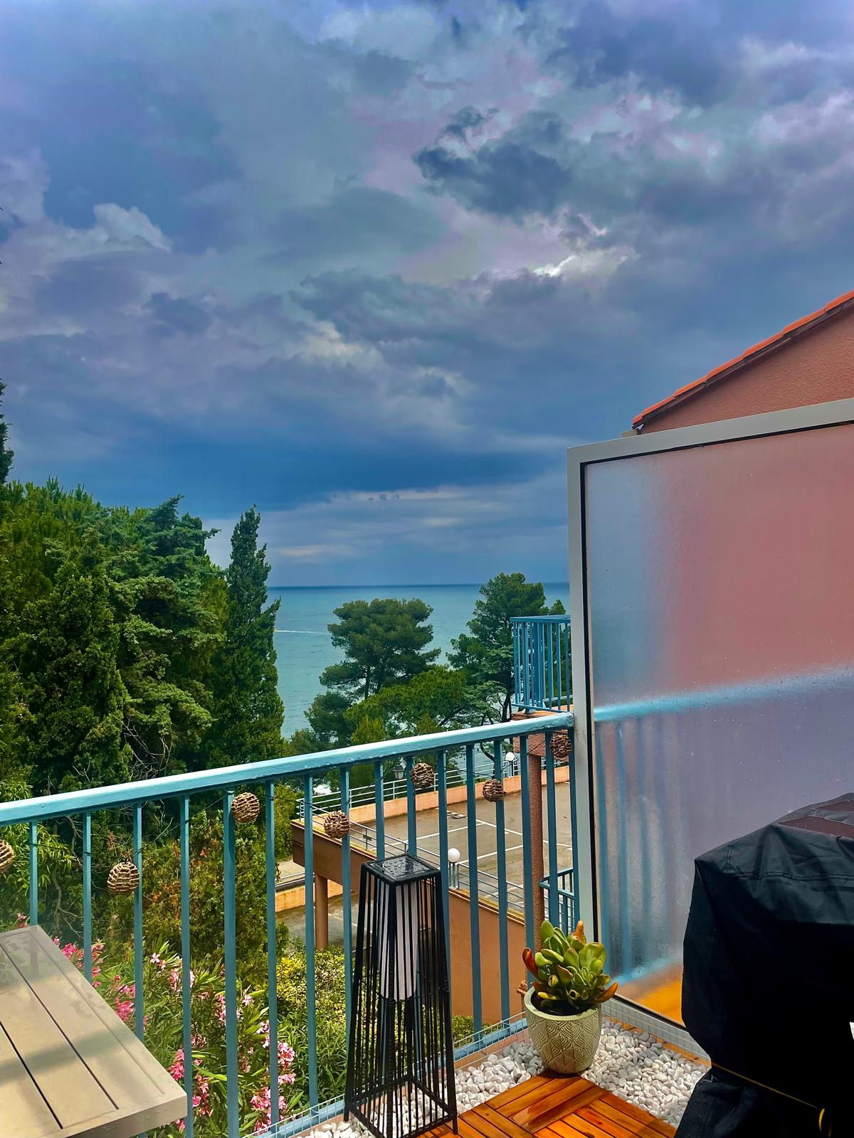 A terrace is presented with a partial view of the sea, framed by lush green shrubs and trees. An outdoor grill and a decorative plant are visible, while the cloudy sky hints at a potential breeze. The atmosphere suggests a serene outdoor dining space.