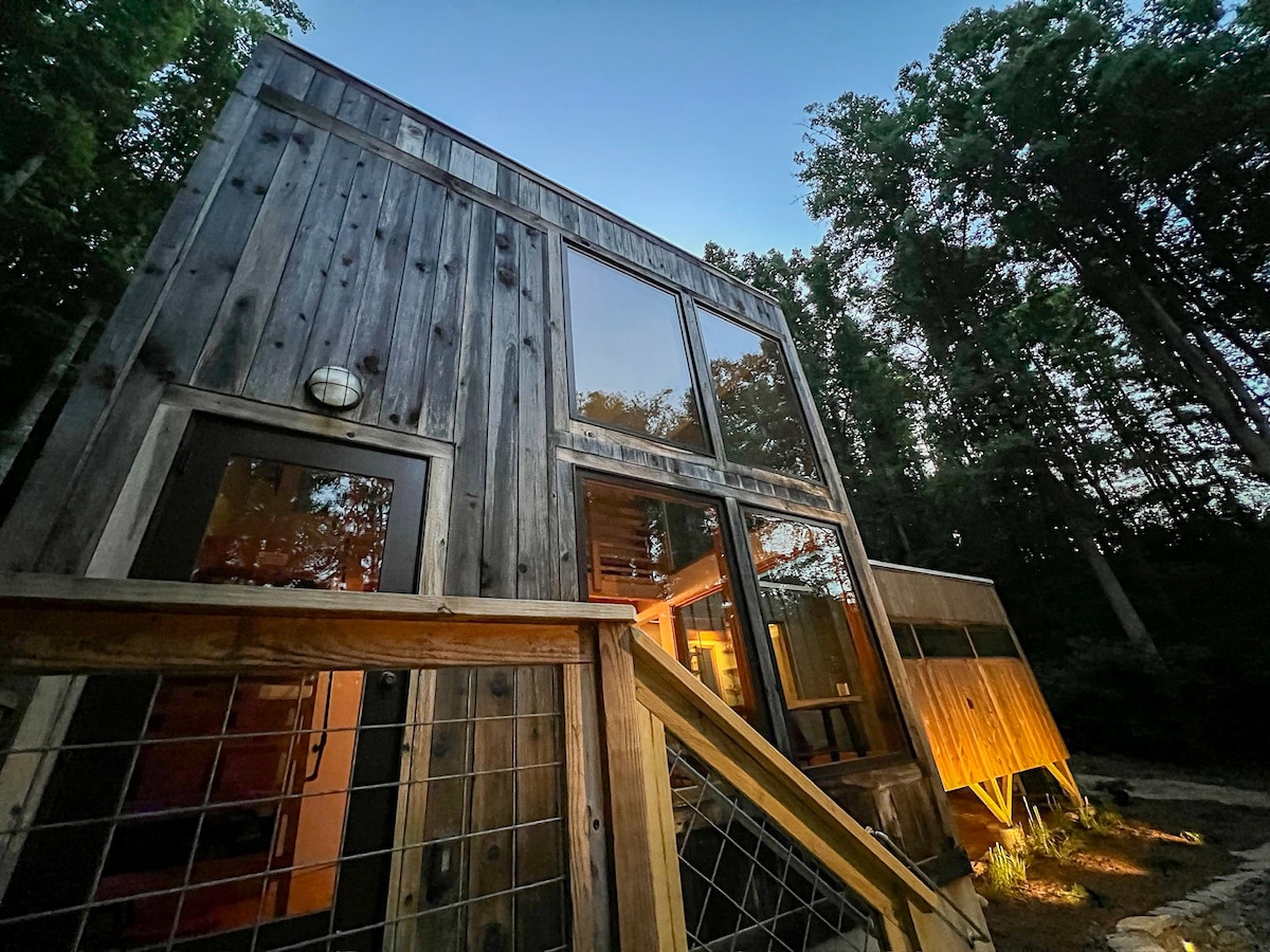 An exterior view of a modern wooden structure is highlighted at twilight, showcasing large glass windows reflecting the towering trees surrounding it. A wooden staircase leads to an entrance, while soft lighting creates a welcoming atmosphere near the entrance and deck area.