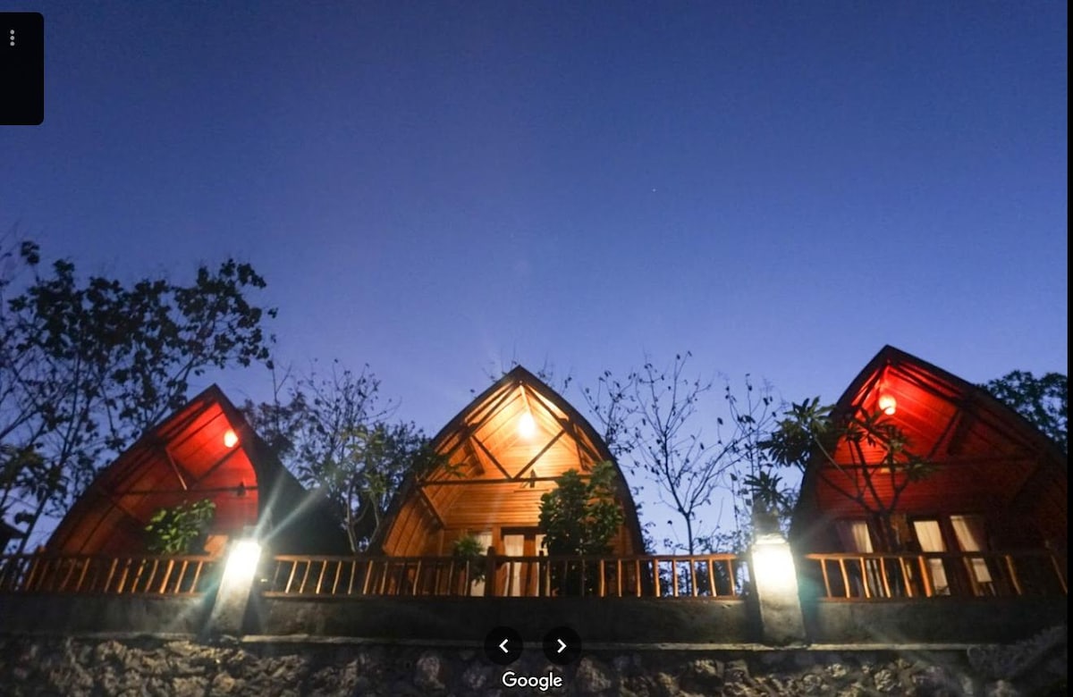 Three rustic cottages are nestled together, featuring distinctive triangular roofs and wooden detailing. Soft lighting highlights their facades, while surrounding greenery creates a natural ambiance. The evening sky transitions from twilight to night, providing a serene backdrop for the structures.
