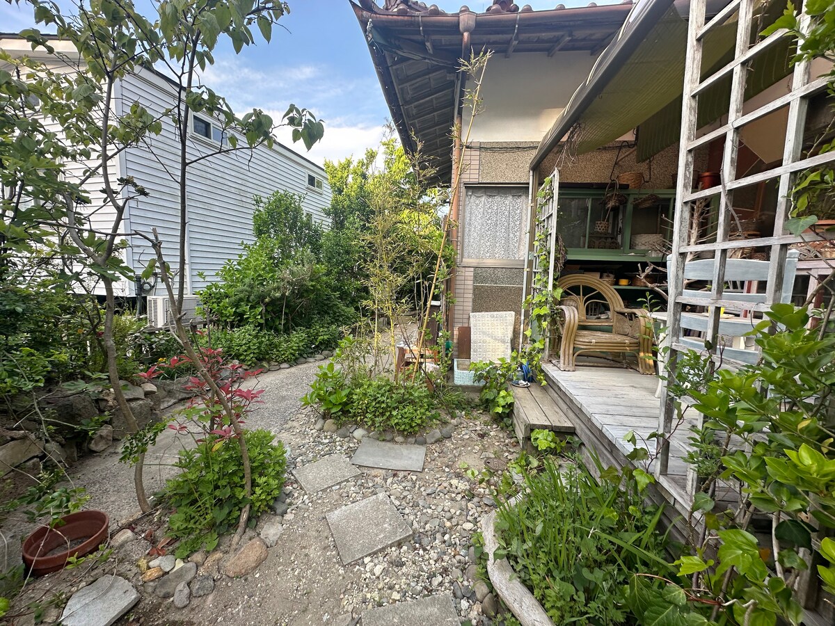 A serene garden path winds through lush greenery, leading to a cozy house. The space is framed by various plants and flowers, with stone tiles guiding visitors. Natural light illuminates the area, creating a peaceful atmosphere for relaxation and enjoyment.