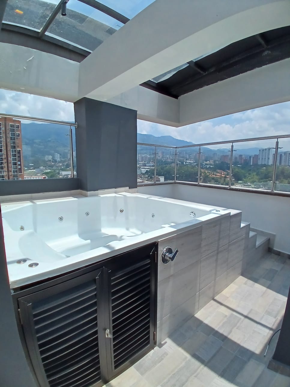 A spacious outdoor terrace features a well-maintained jacuzzi accompanied by seating storage. Stunning views of the surrounding cityscape can be observed through the surrounding glass railing. Bright sunlight filters through the open area, enhancing the inviting atmosphere of the space.