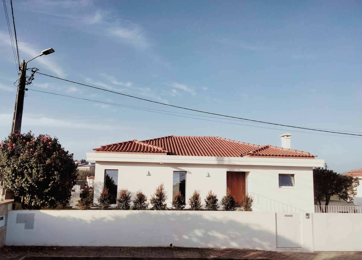A two-story white house with a red-tiled roof is shown against a clear blue sky. Neatly trimmed greenery lines the front of the property, providing a serene and inviting appearance. Simple outdoor lighting fixtures enhance the entryway, maintaining a welcoming exterior.