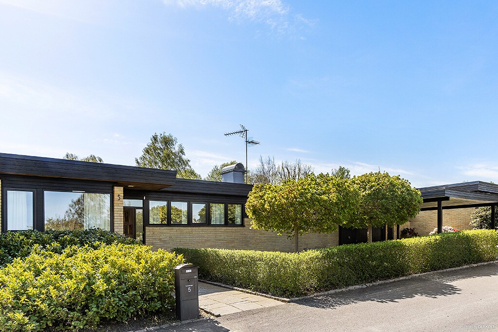 A modern villa is set in a peaceful area, showcasing a sleek exterior framed by well-maintained greenery and low hedges. Large windows provide natural light to the interior, while a paved pathway leads to the entrance. A mailbox stands at the edge of the property.