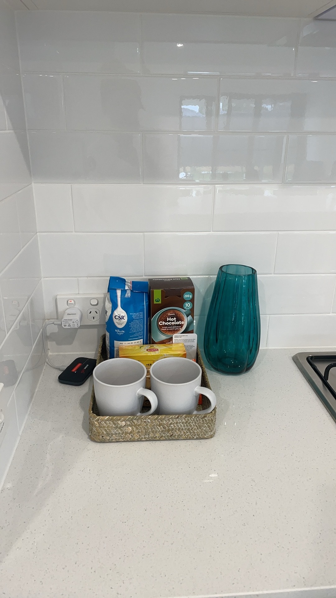 A clean kitchen counter features a collection of two white cups placed on a decorative tray. Nearby, various beverages are neatly arranged, including a carton of milk, a box of tea bags, and a bag of coffee grounds. A blue glass vase adds a pop of color to the setup.