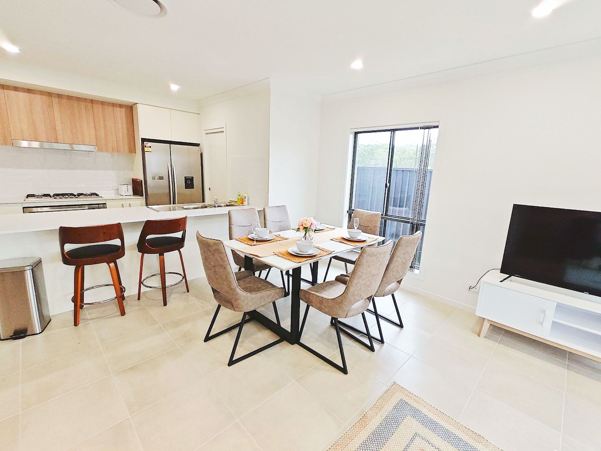 A modern dining area is shown with a sleek black table surrounded by six neutral-toned chairs. The open kitchen is visible in the background, featuring wooden cabinetry and stainless-steel appliances. Natural light enters through a window, enhancing the bright and spacious atmosphere.