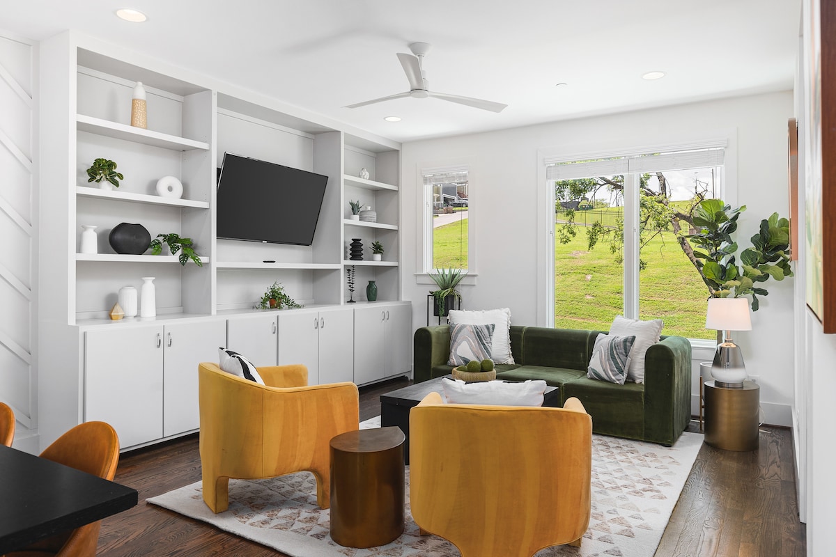 A bright living area features a green sofa paired with two yellow accent chairs, creating a cozy seating arrangement around a dark coffee table. A flat-screen TV is mounted on a white shelving unit, complemented by decorative plants and objects. Natural light streams through the large windows.