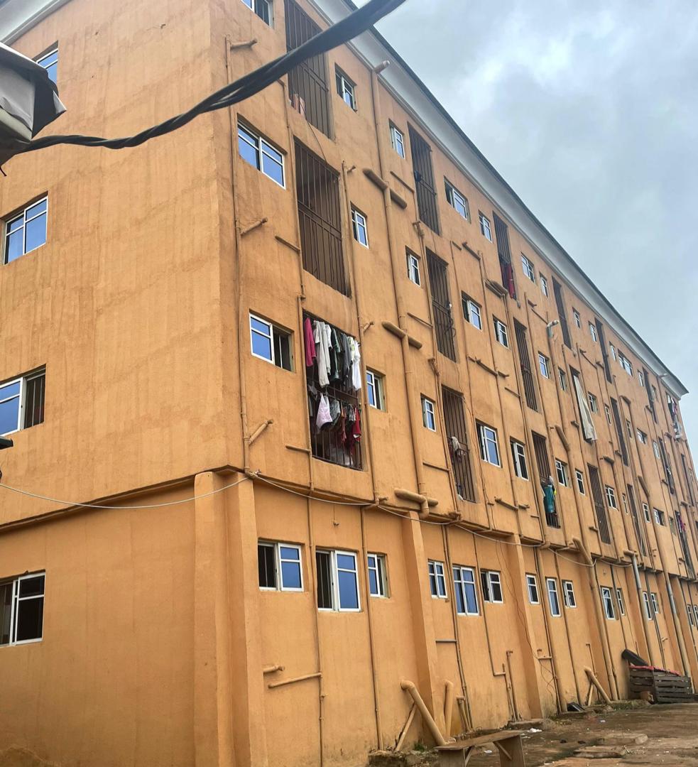 An exterior view of a multi-story building is presented, characterized by its light orange facade. Several windows are visible, with some partially covered by clothing hanging outside. The surroundings include unfinished construction materials, indicating an active development area.