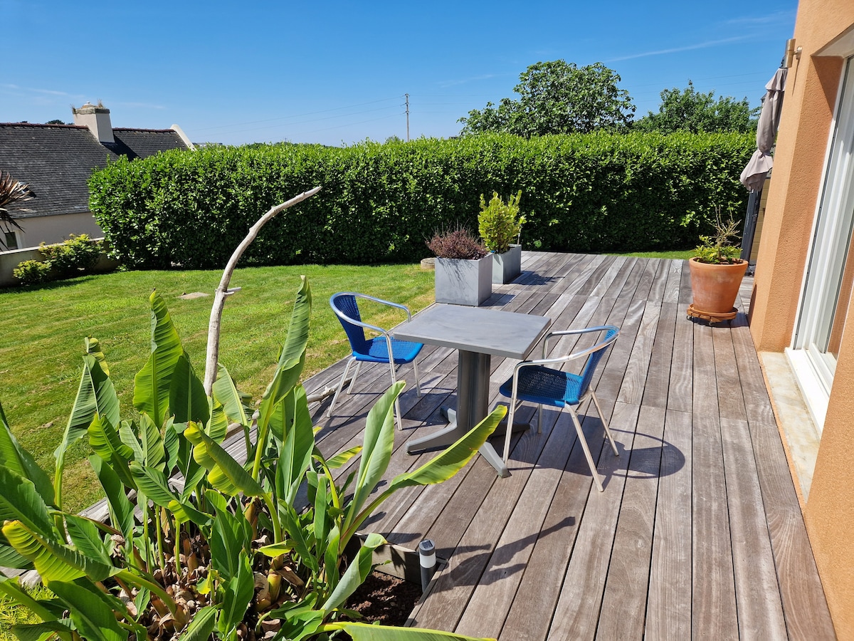An outdoor deck features a rectangular table accompanied by two blue chairs. Lush greenery surrounds the area, including potted plants and shrubs, providing a serene environment. The wooden decking complements the bright sky and sunny atmosphere.
