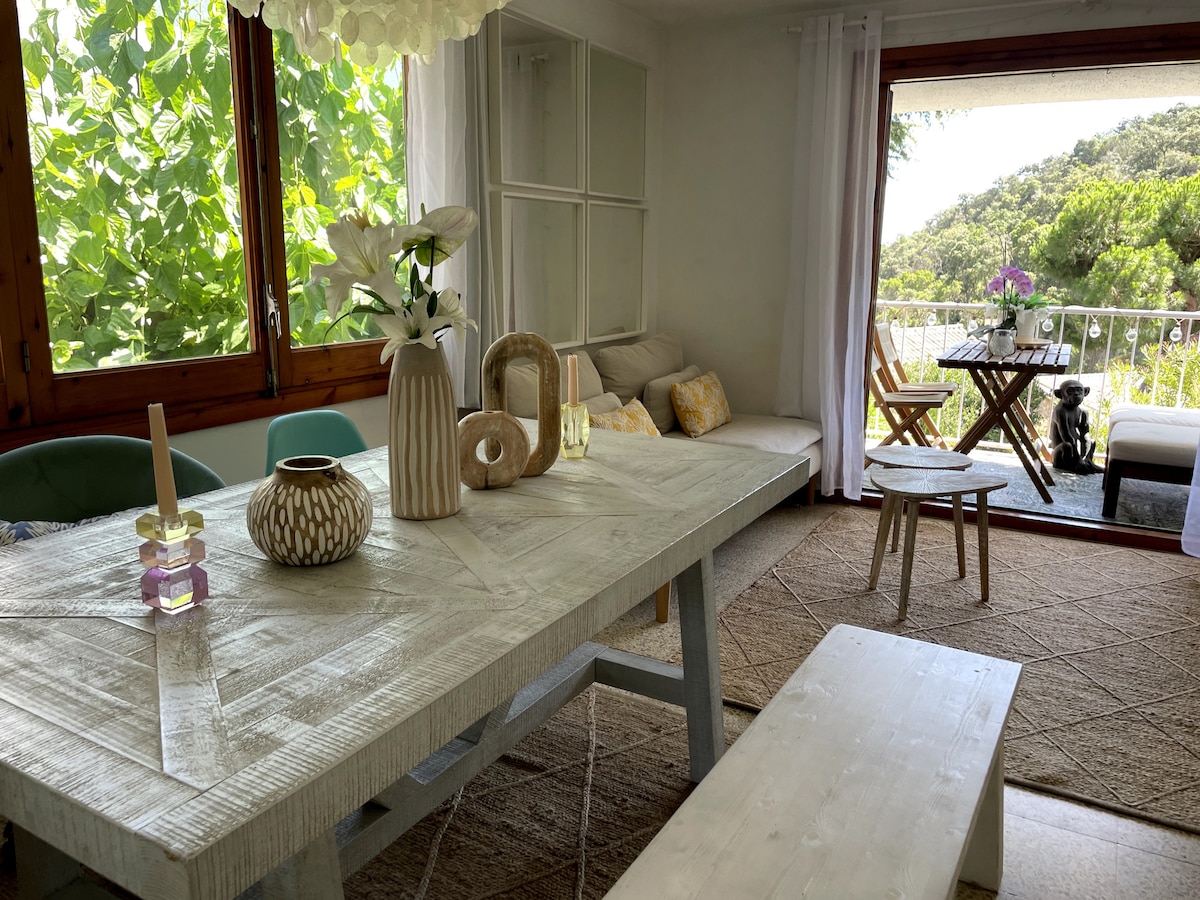 A bright dining and living area features a large, rustic wooden table accompanied by a bench. Natural light enters through expansive glass doors, revealing a peaceful balcony view. Greenery is visible outside, complemented by soft decor accents and a floral arrangement on the table.