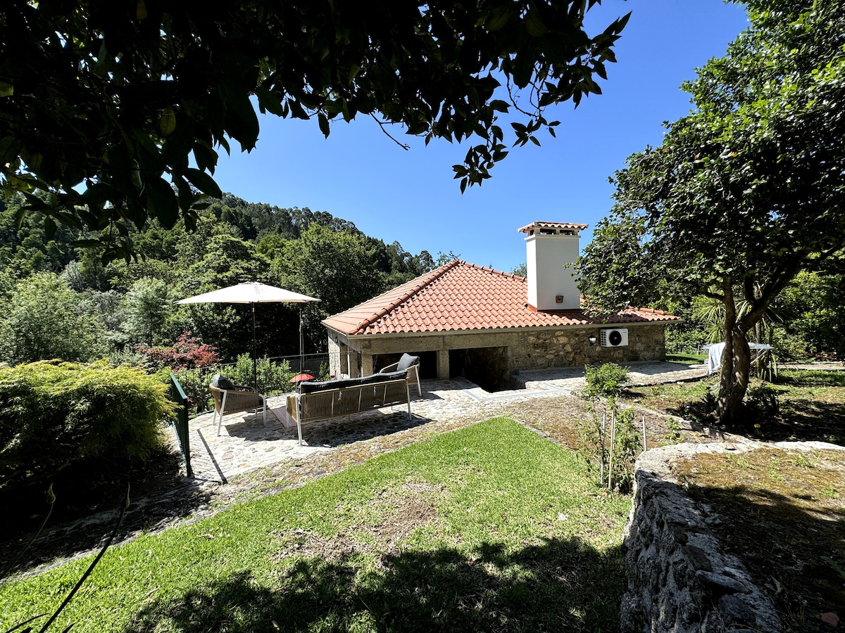 The exterior of a charming rural house is seen, featuring a red-tiled roof and a stone façade. A patio area is visible with outdoor seating and an umbrella, surrounded by lush greenery. The setting is tranquil, blending harmoniously with the natural landscape.