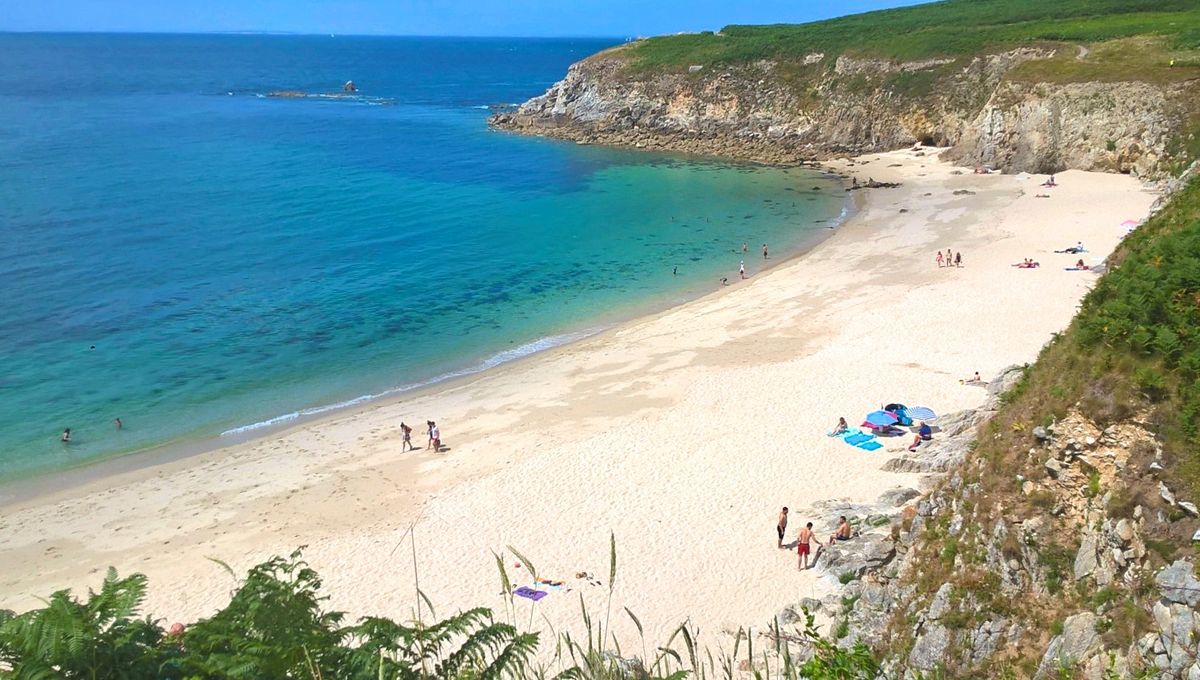 A serene beach is depicted, featuring soft white sand and clear turquoise water. A gentle slope of green foliage leads down to the shoreline, where a few small groups of people are enjoying the sun. The rocky cliffs in the background frame the scene.