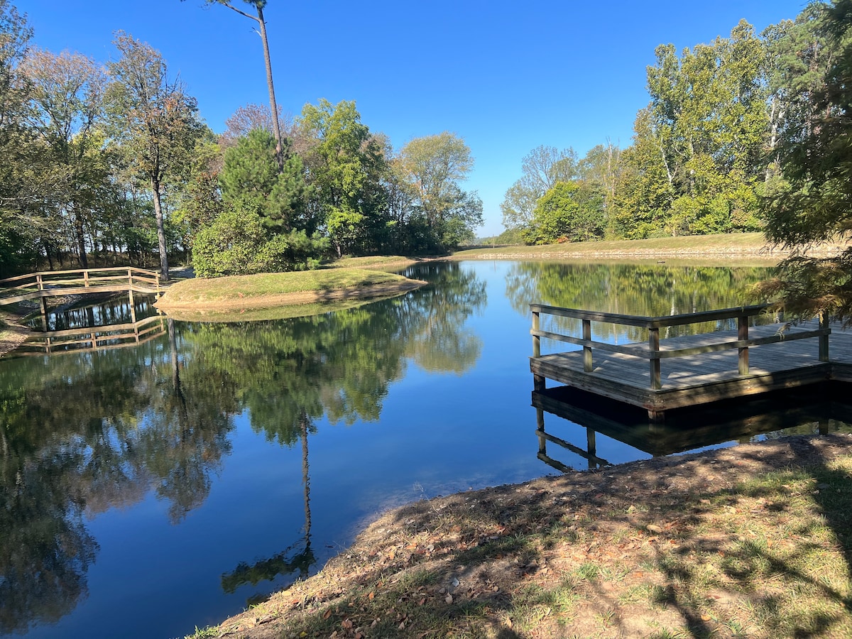 A serene pond is framed by lush greenery, with wooden docks extending gently along the water's edge. Reflections of trees and the clear blue sky can be seen on the calm surface of the water, offering a tranquil setting for relaxation.