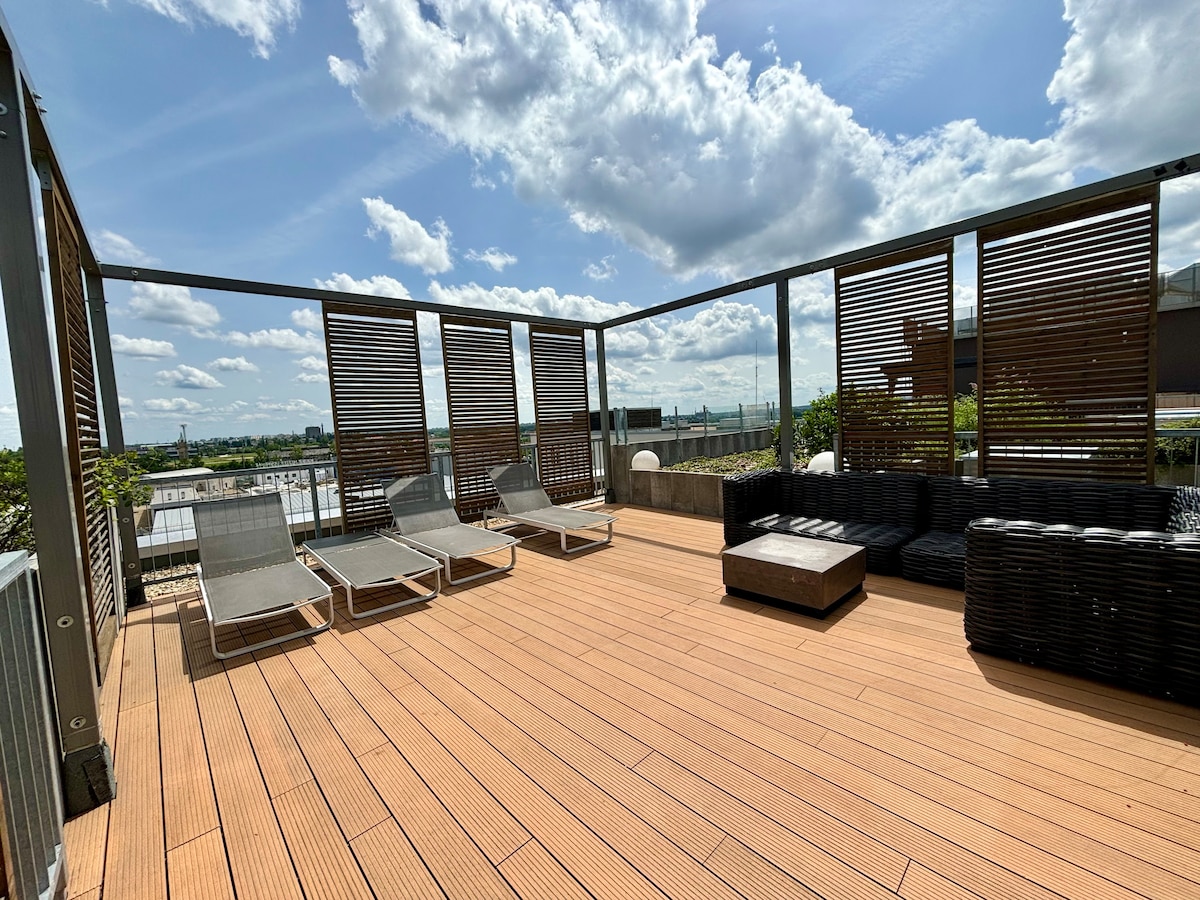 A spacious rooftop garden features wooden decking and a series of lounge chairs for relaxation. A dark wicker seating area is arranged around a low table, while surrounding wooden screens provide partial shade and privacy. The skyline offers scenic views under a bright blue sky.