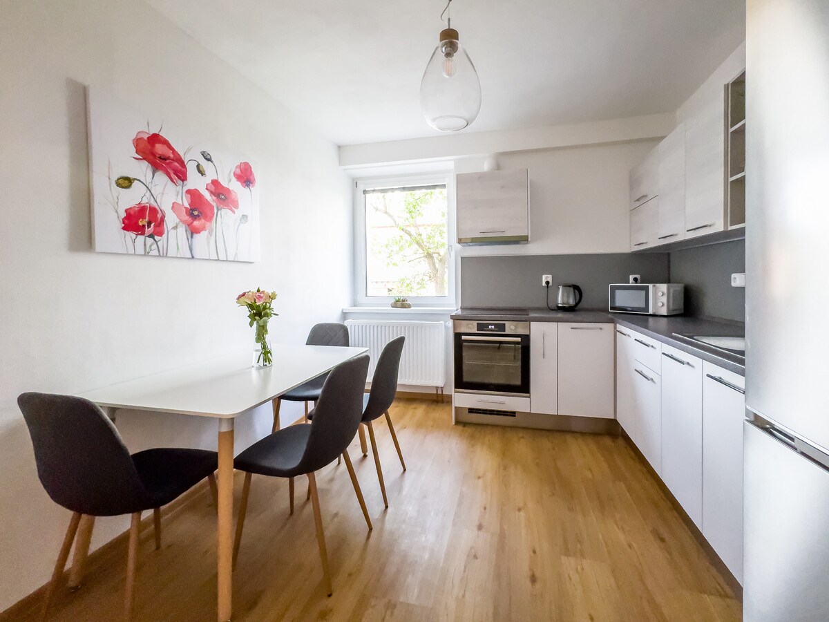 A modern kitchen features sleek white cabinetry and a gray countertop. A dining table with four upholstered chairs is positioned near a window, which allows natural light to enter. A bouquet of flowers adds a touch of color to the space.