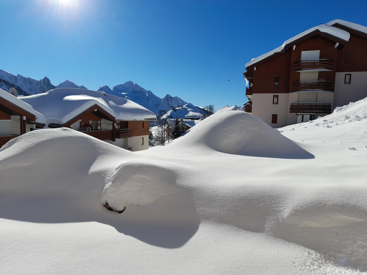 A winter landscape showcases a blanket of snow covering rolling hills and the adjacent buildings. The peaks of distant mountains are visible against a clear blue sky, and the roofs of nearby residences are heavily laden with snow.