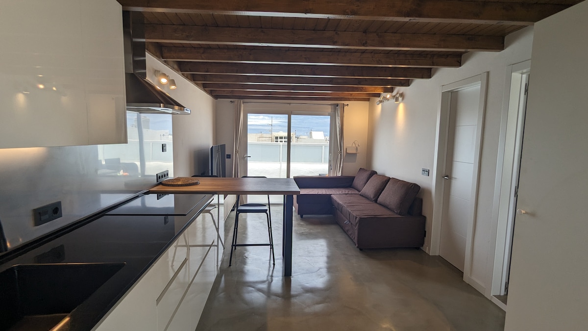 A bright kitchen and living area are presented with polished concrete floors and wooden beams. A comfortable sofa and a dining table are visible, with large windows allowing ample natural light and offering views of the outdoor space.