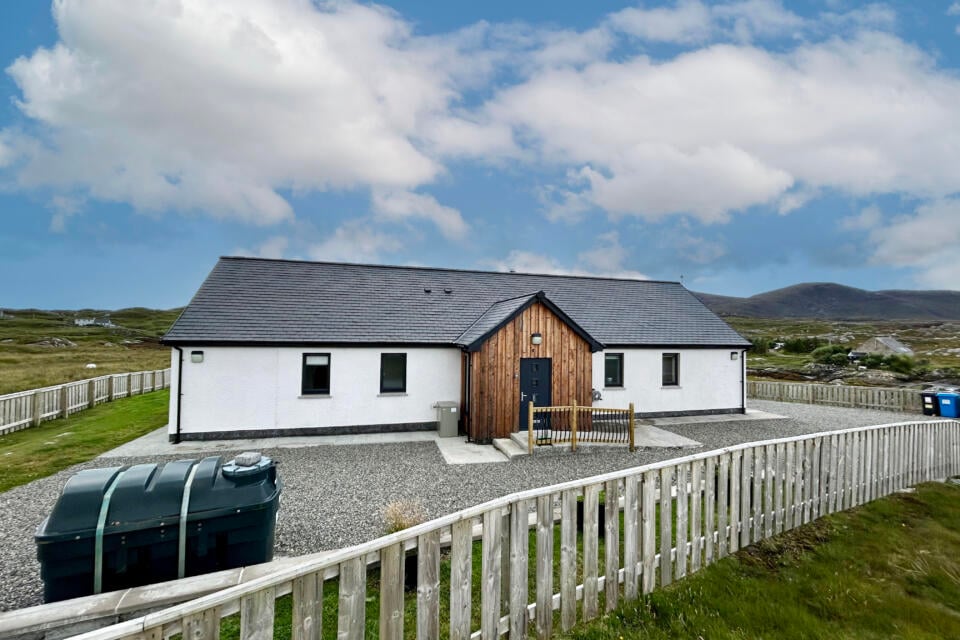 A modern single-story home features a contrasting wooden façade and sleek white exterior. Surrounded by a gravel driveway, the property is enclosed by a low wooden fence, with rolling hills and a luminous sky visible in the background.