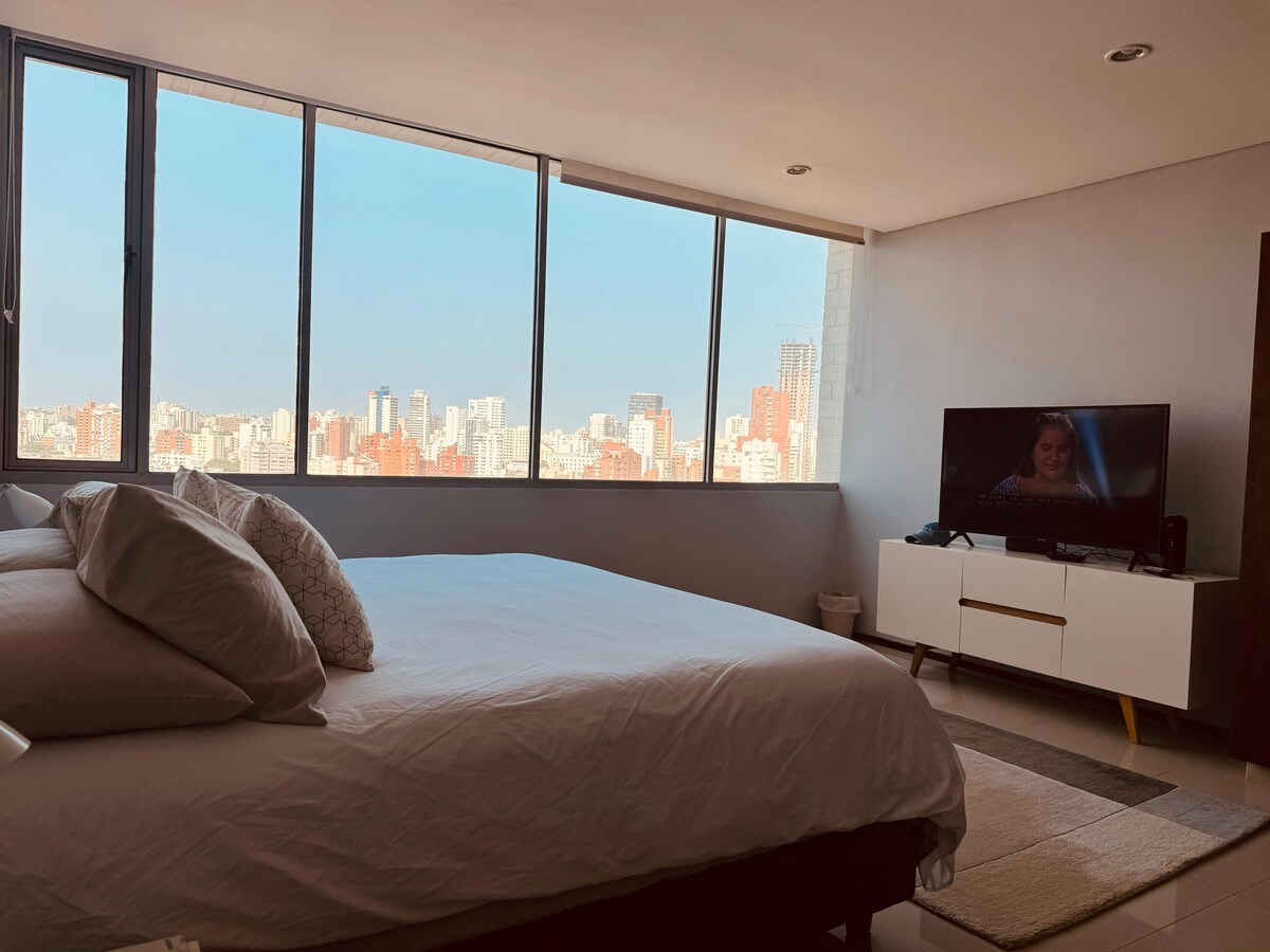 A bright bedroom features a spacious bed with multiple pillows, complemented by a large window offering expansive views of the city skyline. A modern TV is positioned on a sleek console. Soft natural light floods the room, enhancing its inviting atmosphere.