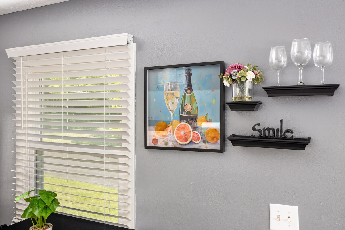 A wall adorned with a framed painting featuring a champagne bottle and fresh fruits is complemented by two black shelves holding three glasses and a small bouquet of flowers. A window with white blinds allows natural light to filter in, enhancing the cheerful atmosphere.