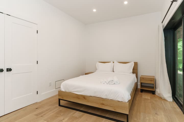 غرفة نوم بسيطة مع سرير أبيض وباب أبيض وستارة بيضاء. السرير مصنوع من ملاءات ووسائد بيضاء، ومنضدة خشبية مع مصباح بجانبه. الغرفة مشرقة ومتجددة الهواء، مع نوافذ كبيرة تسمح بدخول الضوء الطبيعي.