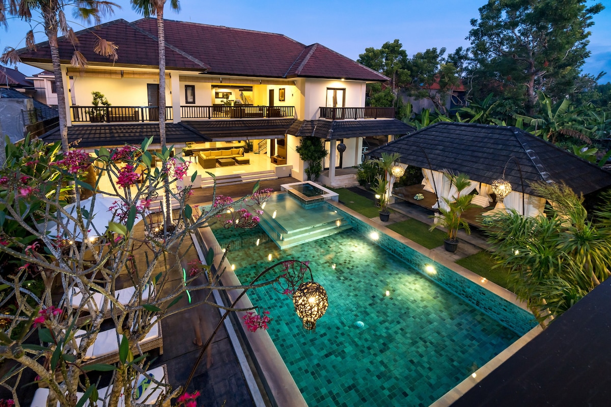 An aerial view highlights the spacious villa surrounded by lush greenery. A large swimming pool reflects soft lighting, with decorative lanterns enhancing the atmosphere. The two-story structure showcases balconies and expansive windows, seamlessly integrating indoor and outdoor living spaces.
