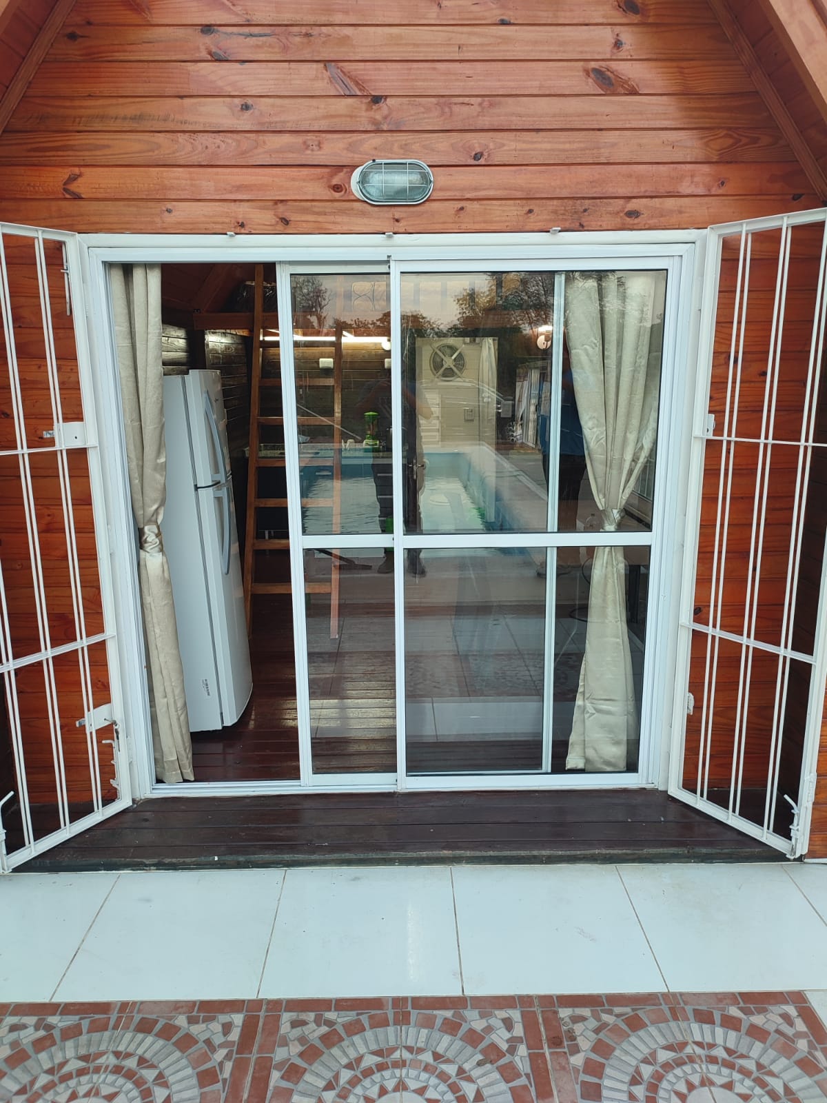 Double glass doors open outward, framed by white metal bars, revealing a glimpse of an inviting interior with wooden accents. Light beige curtains drape beside the doors, and a modern fridge is visible through the entrance, complementing the warm wooden exterior.