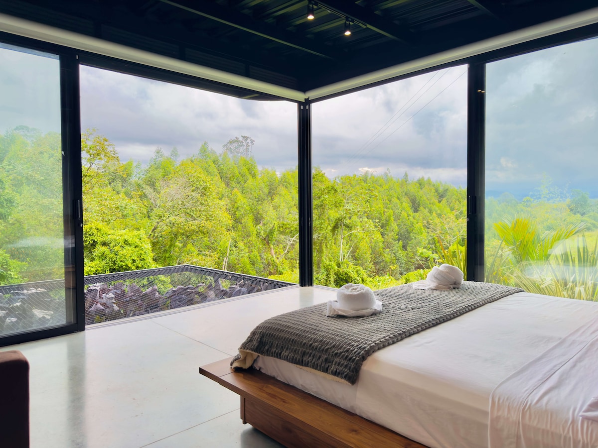 A serene bedroom is featured with a spacious bed adorned with a neutral-colored blanket. Large glass walls provide panoramic views of lush greenery outside. The room is illuminated by natural light, enhancing the tranquil ambiance. Two neatly placed towels add a welcoming touch.