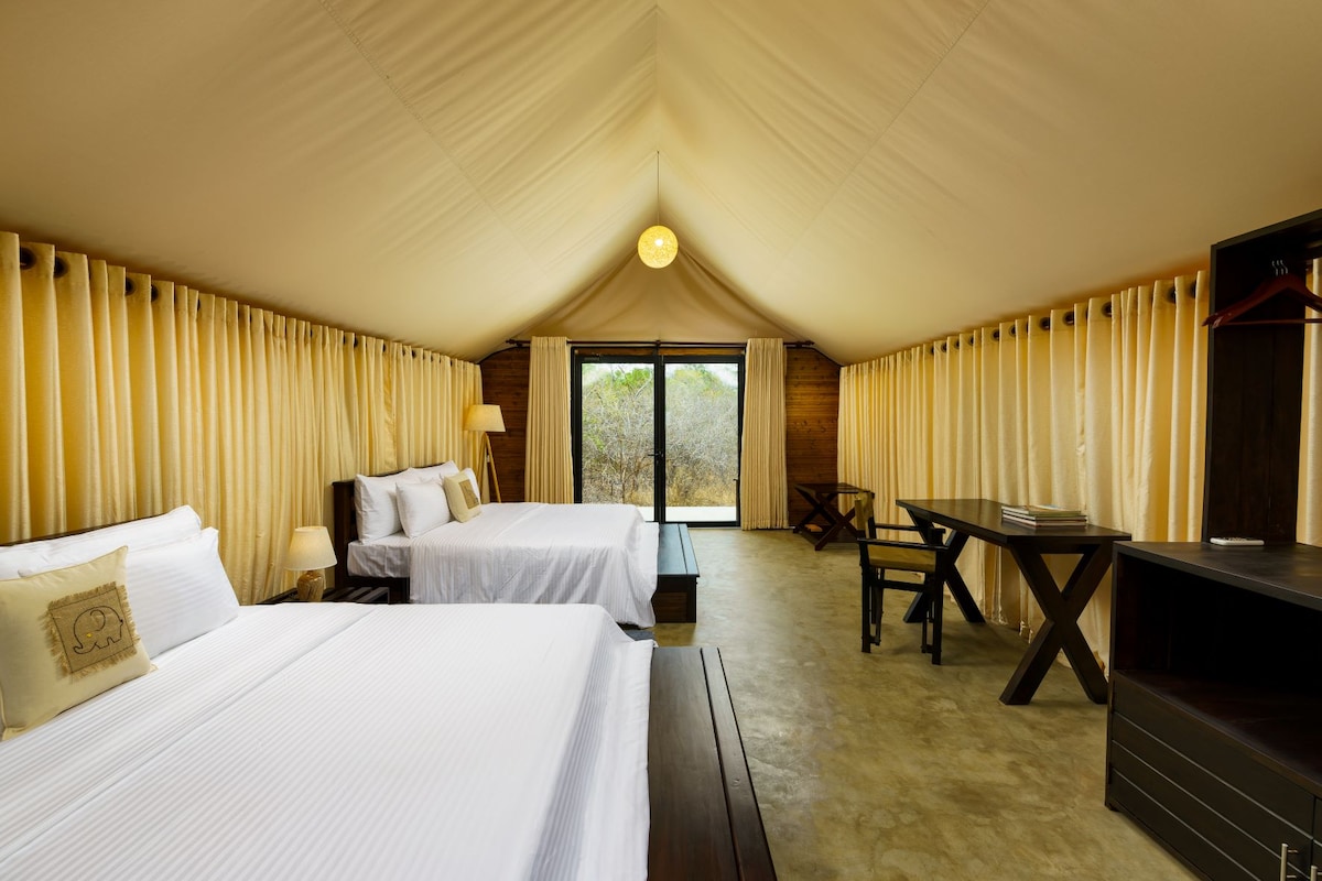 The spacious accommodation features two beds, each adorned with crisp white linens. A large window offers views of the surrounding landscape, while soft curtains frame the view. A wooden desk and chair provide a workspace, complemented by warm lighting and natural textures.