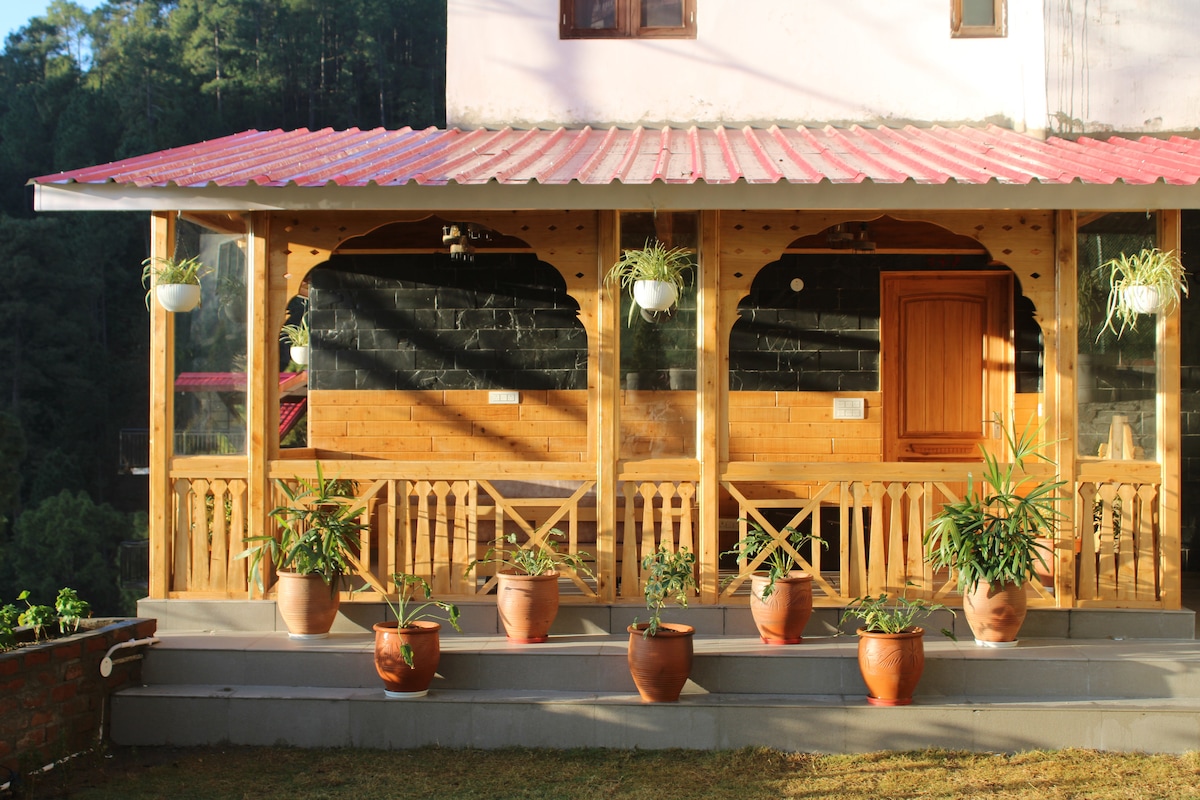 A welcoming outdoor sitting area features a covered wooden structure with a red roof and natural wood finishes. Potted plants line the steps, bringing greenery to the space. The area offers views of the surrounding nature and is designed for relaxation.