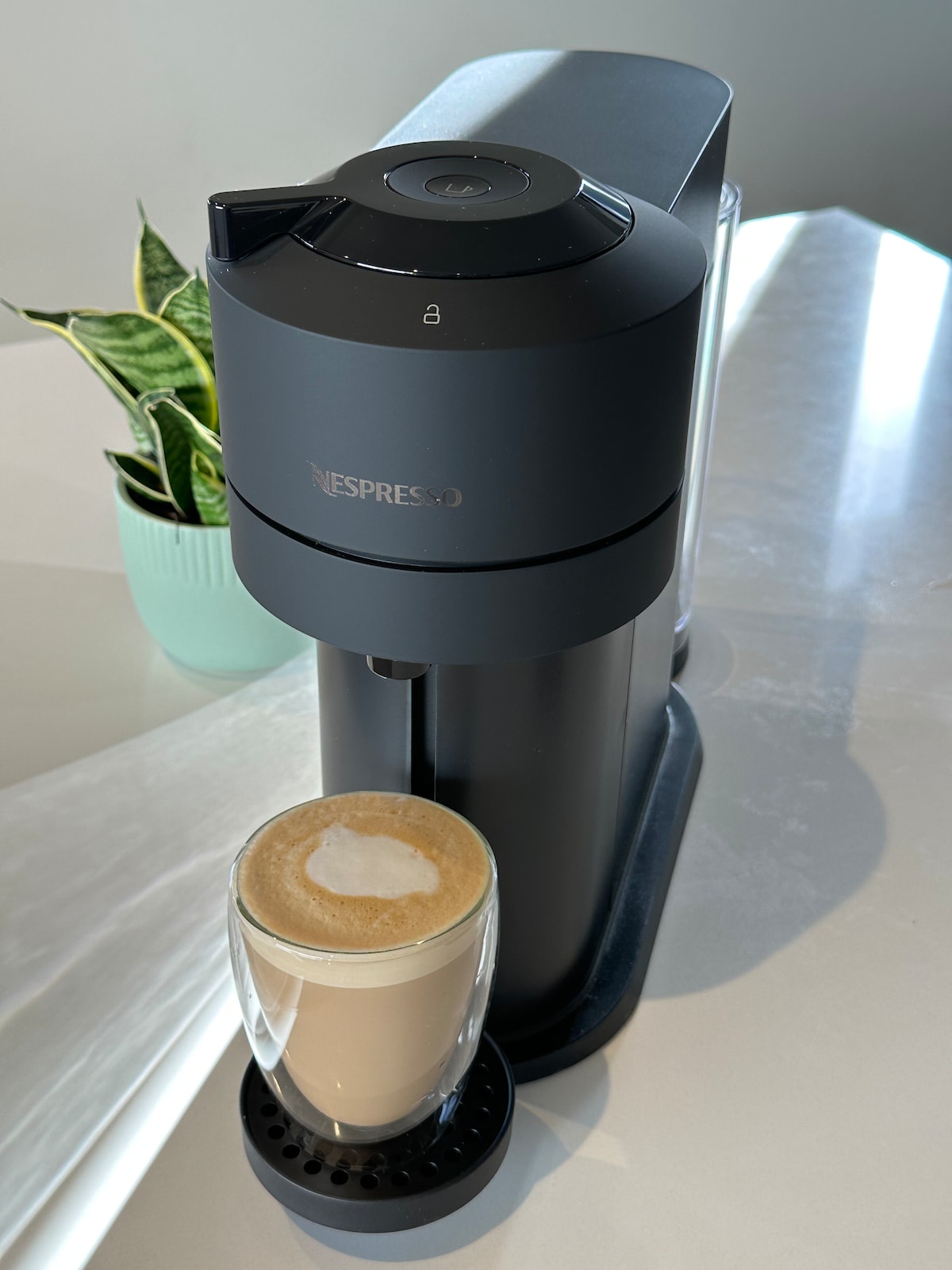 An Nespresso coffee machine is displayed on a sleek countertop, positioned next to a small potted plant. A glass filled with creamy coffee sits in the machine's cup holder, showcasing a delicate frothy topping.