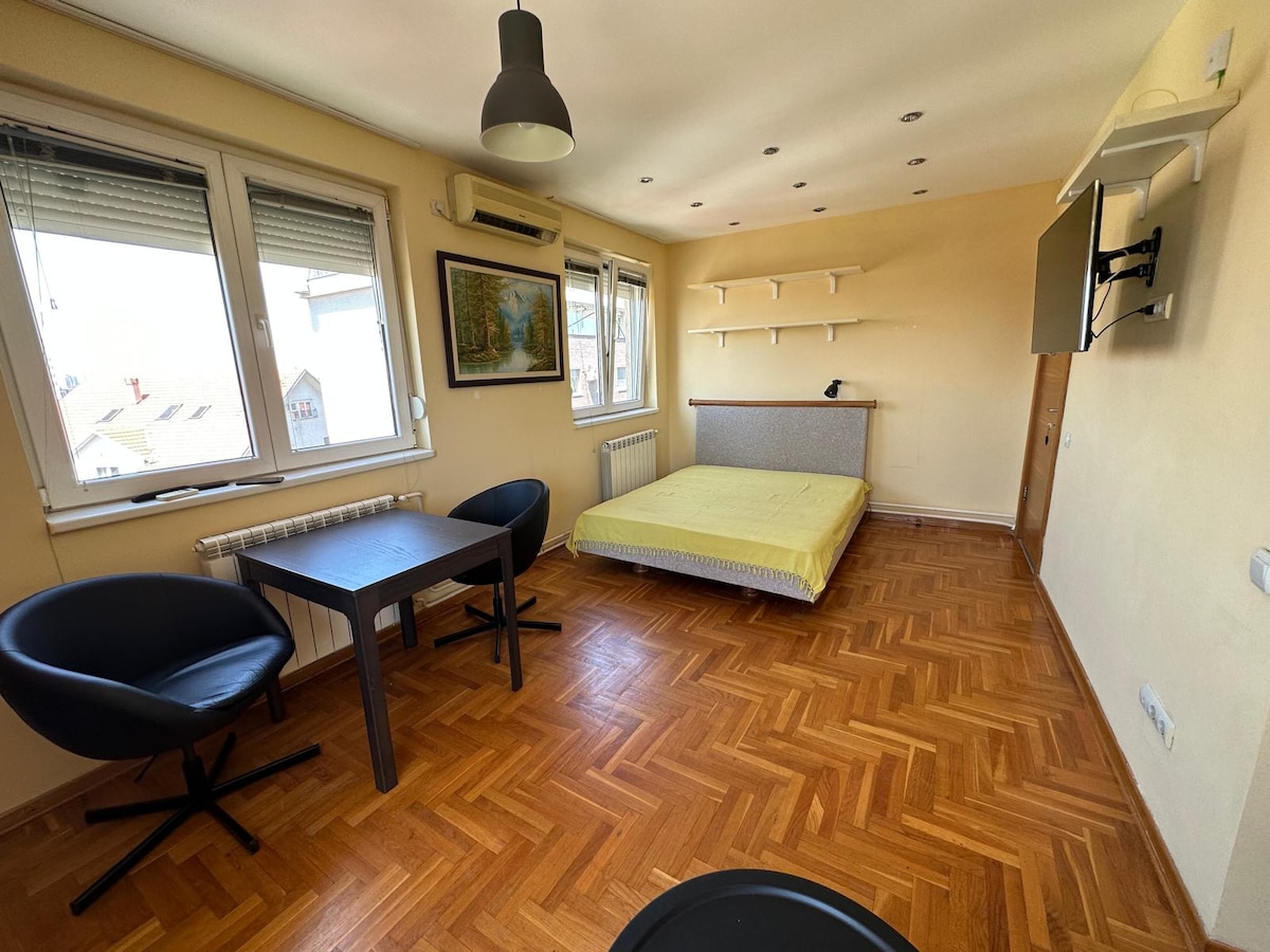 A bright room features a spacious bed against a wall, accompanied by a small dining table with two chairs. Natural light enters through large windows with shades, and a mounted television is visible on the wall. The herringbone-patterned wood flooring adds warmth.