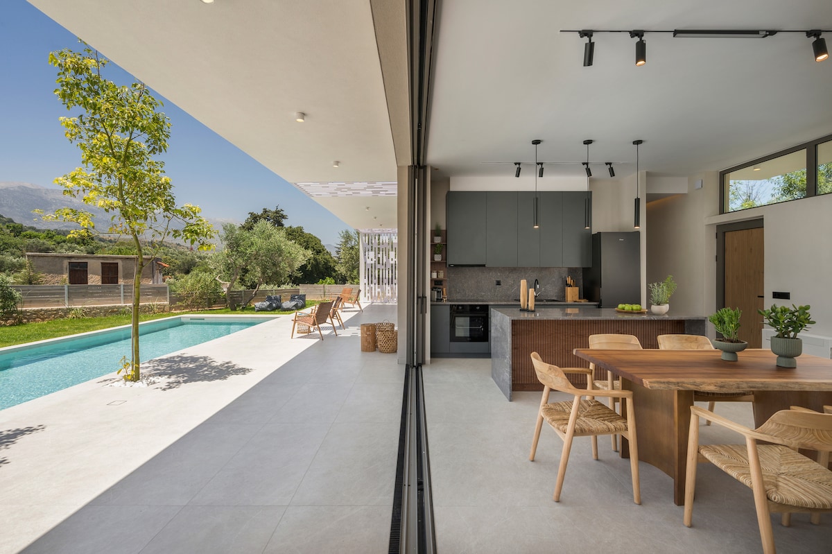 An open-plan living space features a dining area with a wooden table surrounded by woven chairs. The kitchen showcases modern cabinetry and appliances. Large glass doors provide seamless access to the outdoor pool area, where wooden lounge chairs are positioned for relaxation.