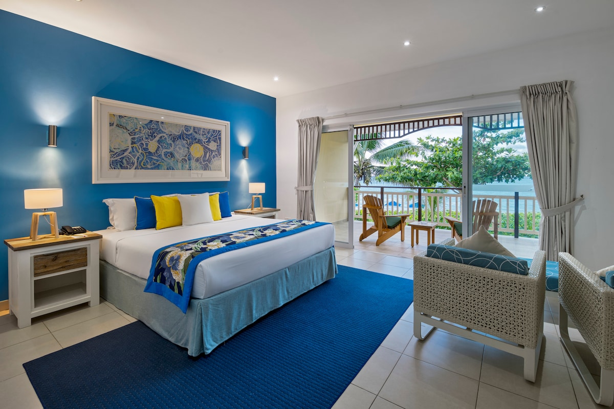 A spacious room features a large bed with decorative pillows, complemented by a vibrant blue accent wall. Natural light fills the area through sliding glass doors that open to a balcony with outdoor seating, overlooking lush greenery and the nearby beach.