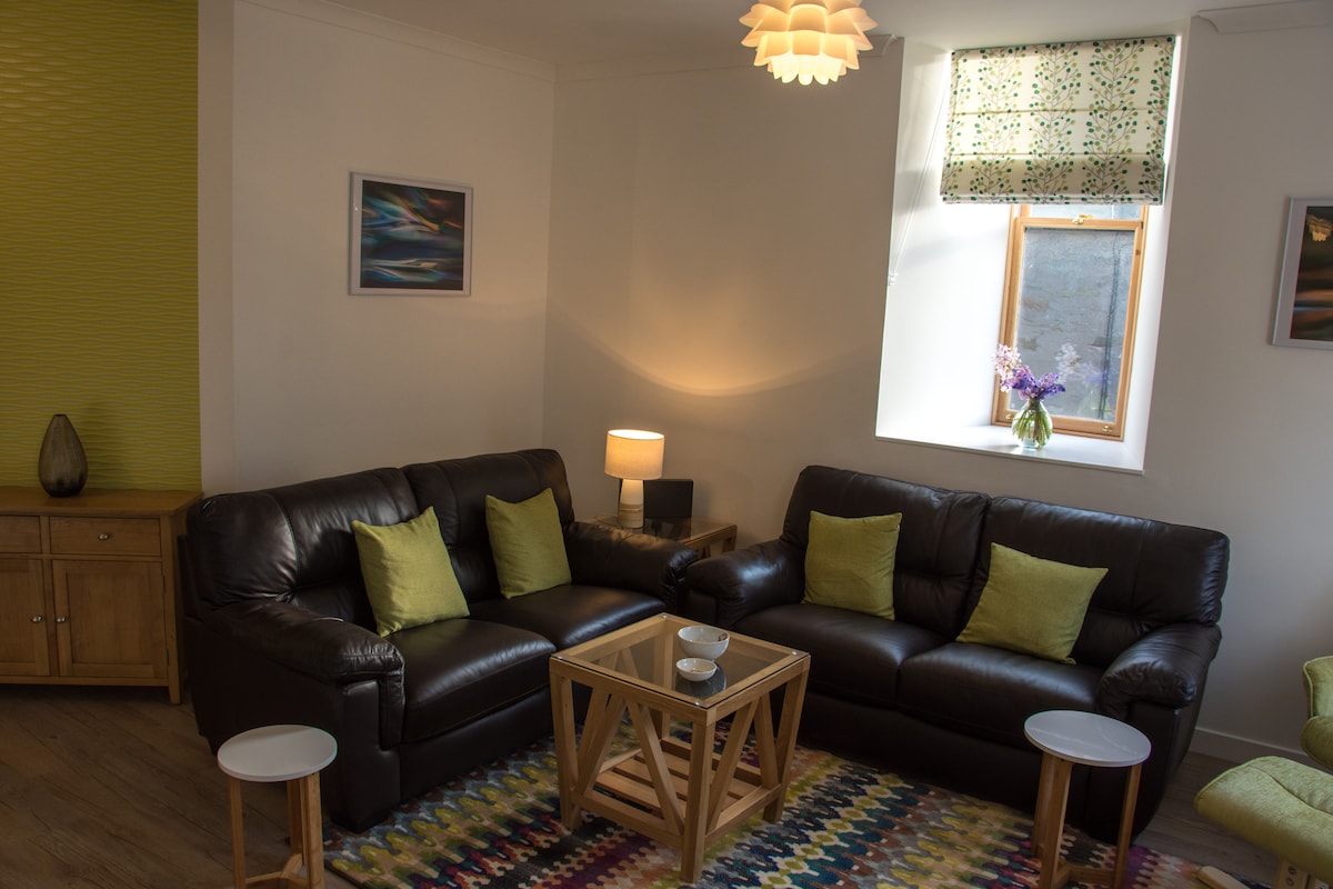 A cozy living area features two dark leather sofas with green accent cushions, positioned around a wooden coffee table. A small light illuminates the space, while a window dressed in patterned fabric allows natural light to enhance the room's inviting ambiance.