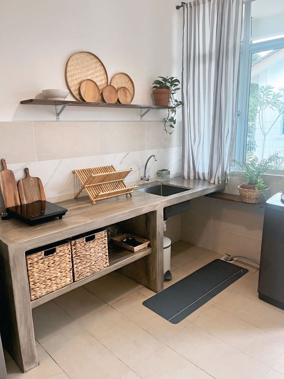 A well-organized kitchen area features a sleek counter with a double sink and modern fixtures. Above, various dishware is neatly arranged on a shelf. A small potted plant adds a touch of greenery, while wicker baskets provide storage underneath. Natural light filters in through a window.