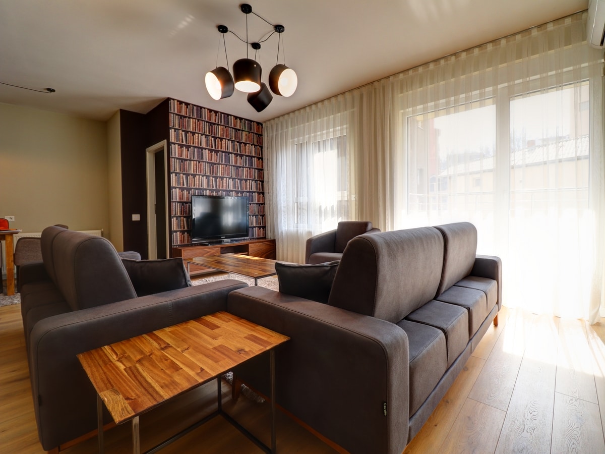 A spacious living area features multiple comfortable sofas arranged around a wooden coffee table. Natural light fills the room through large windows adorned with sheer curtains, while a patterned wall of books adds character. A television is positioned for easy viewing.