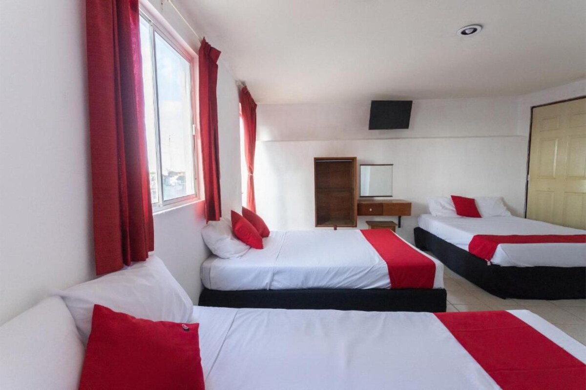 The room features two spacious beds, each adorned with crisp white linens and red accent pillows. A window allows natural light to fill the space, complemented by white walls and red curtains. A flat-screen TV and a wooden dresser add to the functionality.