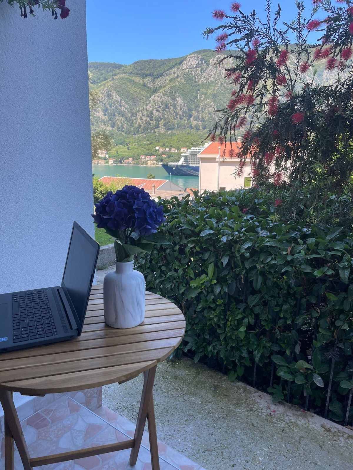 A small round table sits on a patio, accompanied by a laptop and a vase of blue hydrangeas. Lush green foliage borders the area, with views of mountains and a body of water in the background under clear skies.