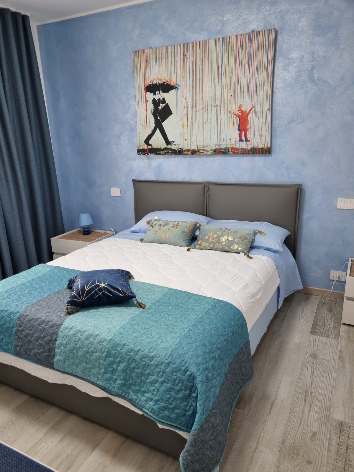A serene sleeping area features a spacious bed with a light blue and patterned quilt. Decorative pillows add a touch of color, and a vibrant artwork hangs above the headboard. Soft curtains frame the window, allowing natural light to enhance the calming blue walls.