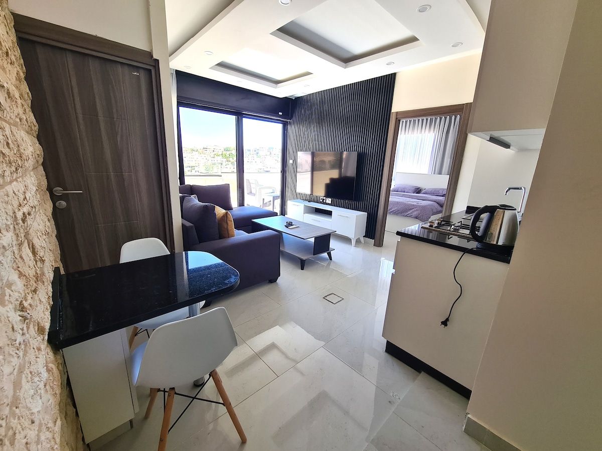 An open-plan living area features a modern design with natural stone accents and large windows. A comfortable seating arrangement includes a sofa and coffee table, while a dining table sits nearby. The kitchen is equipped with essential appliances, creating a seamless space for relaxation and dining.
