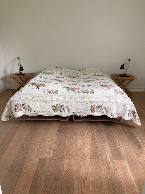 A spacious bed is presented with a floral-patterned quilt. Two bedside tables with lamps sit on either side, enhancing the simplistic decor. The hardwood floor reflects a warm tone, contributing to a calming ambiance in the room.