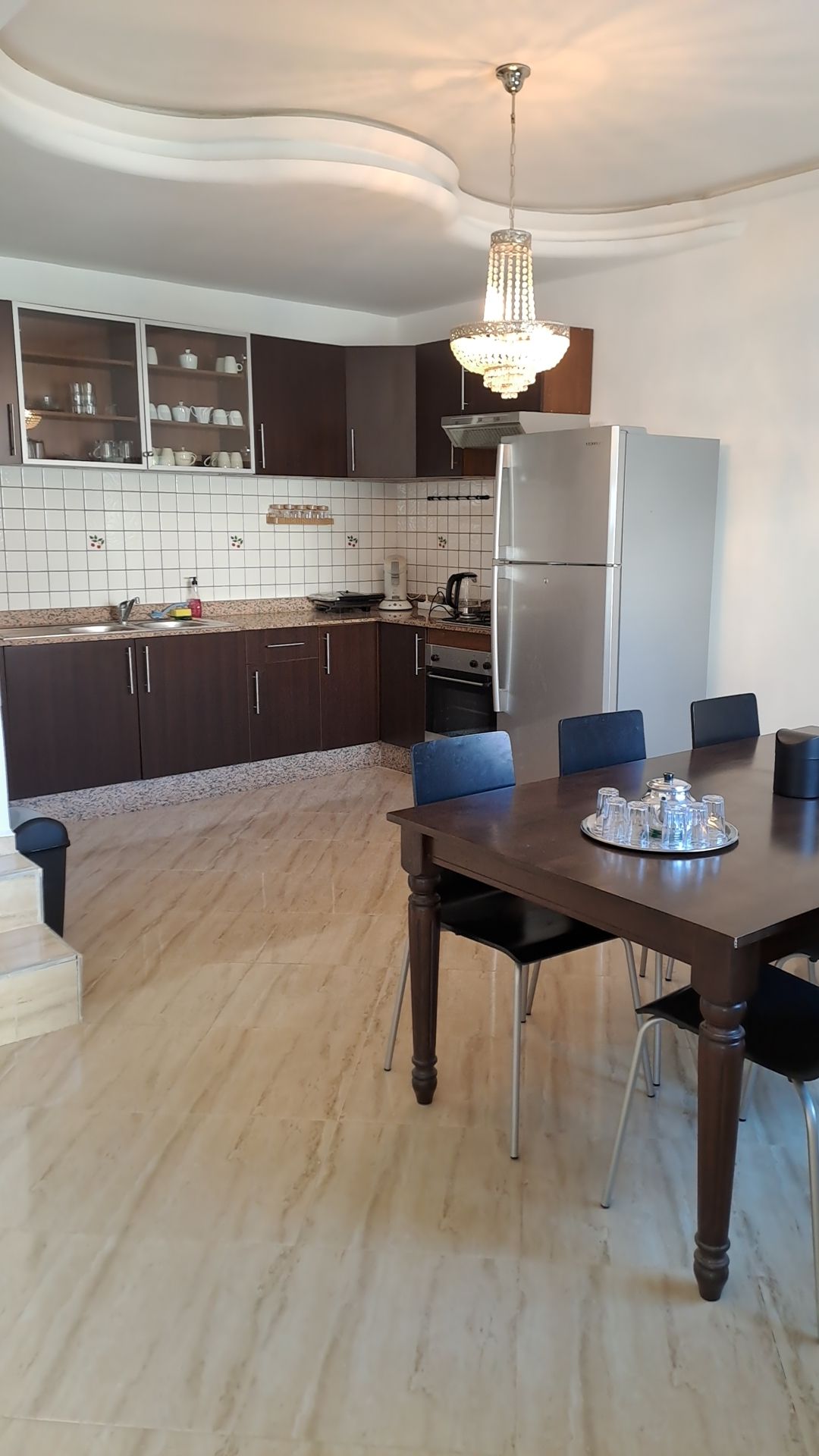 A modern kitchen area features dark cabinetry and a light-colored tiled backsplash. A dining table with four black chairs is positioned nearby. Stainless steel appliances, including a refrigerator and stovetop, are visible, along with a bright overhead light fixture.