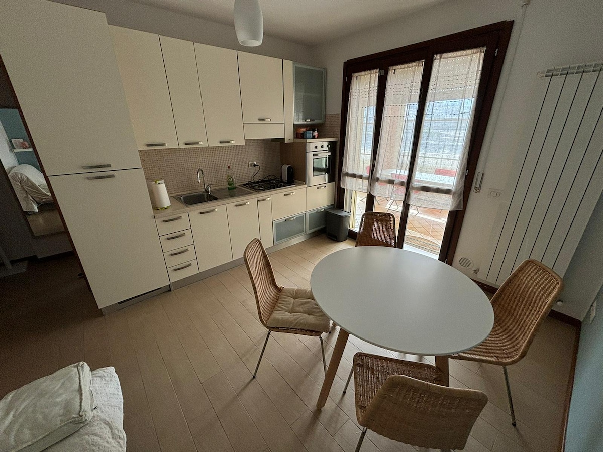 A compact kitchen area features light cabinetry and modern appliances. A circular dining table is surrounded by four woven chairs. Large windows provide natural light, and a door leads to the furnished balcony, enhancing the space's overall functionality.