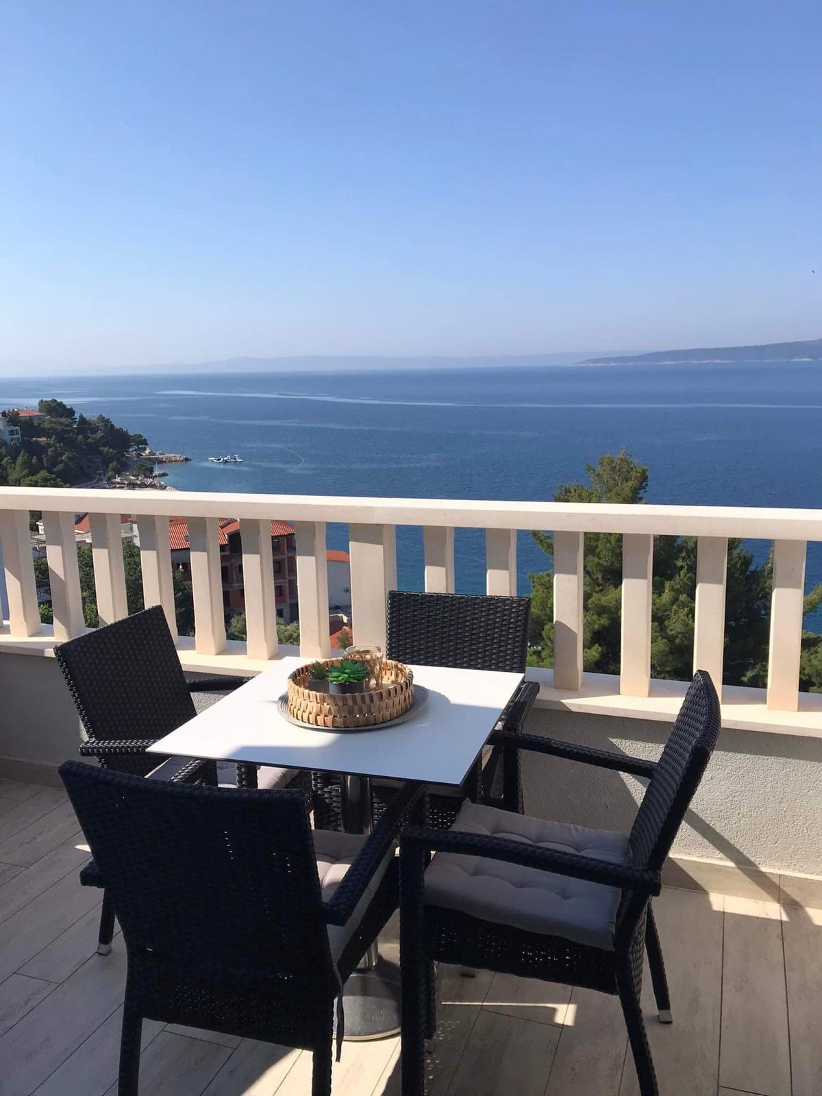 A balcony is presented with a table and four chairs, offering a clear view of the sea and surrounding landscape. A woven basket is placed on the table, complementing the outdoor setting. The horizon and water reflect bright blue tones under a clear sky.