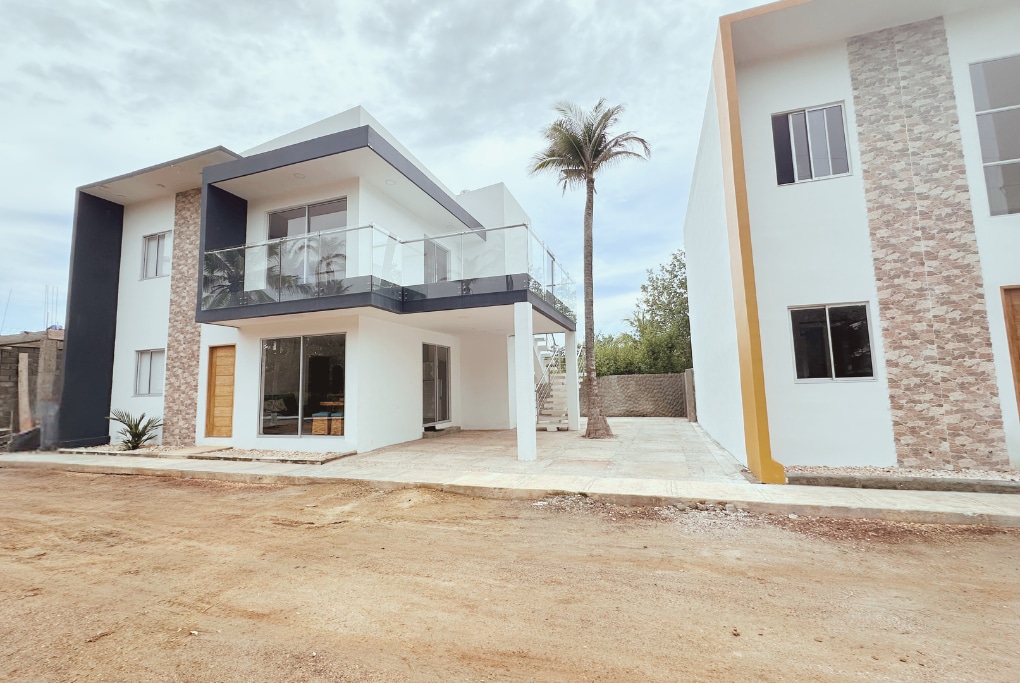 The exterior of a two-story house is showcased, featuring a sleek modern design. A balcony with glass railings offers views of the surroundings. Palm trees and a gravel pathway lead to the entrance, enhancing the inviting atmosphere of the property.