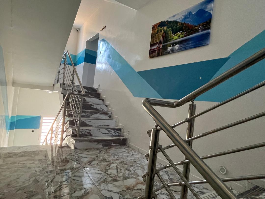 A modern staircase is depicted, featuring a polished marble floor and sleek stainless steel railing. The walls are painted in calming blue and white stripes, complemented by a landscape painting. Natural light enters from the adjacent windows, enhancing the spacious feel.