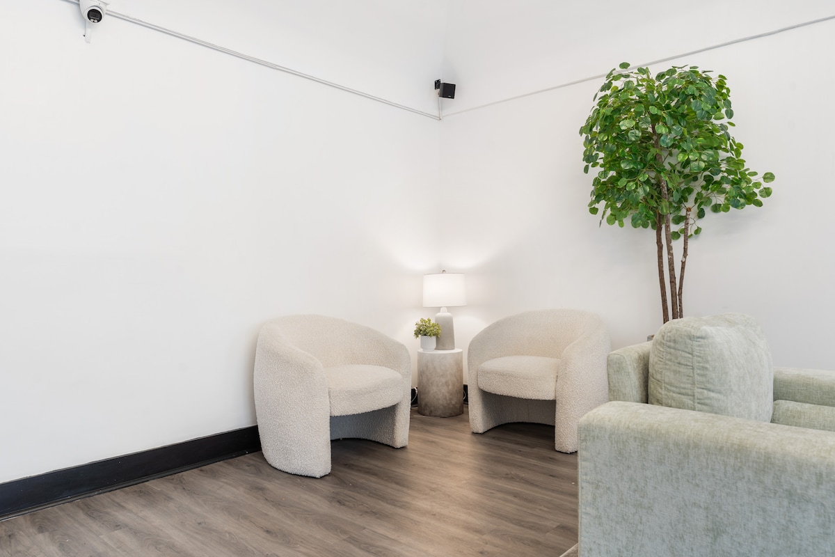 A cozy shared lobby area features textured cream-colored chairs paired with a small side table. Soft lighting from a nearby lamp illuminates the space, while a leafy plant adds a touch of greenery, enhancing the inviting atmosphere for guests.